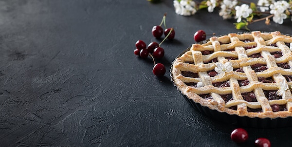 A homemade cherry pie with whole cherries next to it and cherry blossoms in the background.